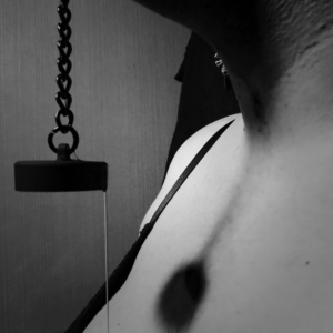 A close-up in black and white of spit dripping in a thread from the chained stopper of the gag.