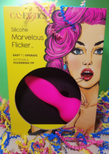 A yellow box standing against a green background with multicolor confetti scattered about. Art of a pink-haired femme with her mouth agape overshadows a cut-out through which a pink tongue-shaped vibrator is visible. The plastic for the cut-out has been removed.