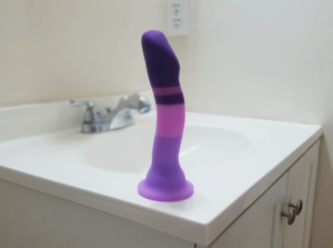 An enticingly but mildly curvaceous dildo sitting on a white bathroom sink counter. The dildo is cleanly striped with four different shades of purple and has a suction cup.