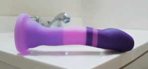 The Avant D2 Purple Rain Dildo laying on its side on the sink counter, showing off its subtly curved body, which ends with a squatter base and broad suction cup.