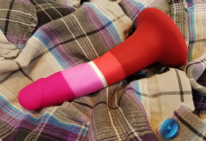 The P3 sprawled on a flannel shirt. Its head is a dark pink; the shaft is striped with a link pink, then a thin strip of white, then pink-red; the base of the shaft and suction cup is dark red.