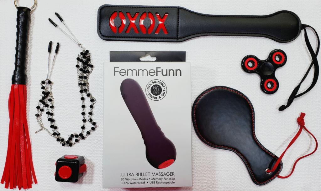 A FemmeFunn box laying among red and black kink products (including Jeweled Nipple Clamps and the XOXO Paddle) and fidget toys. The vibrator on the box is baseball bat shaped and has a bulbous base with a large, single button.