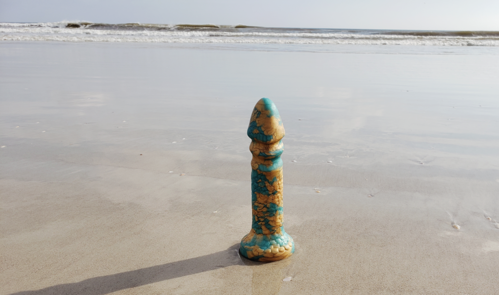 The Aqua-King standing upright on the beach with the ocean far off in the background. The sun has created a long shadow of the dildo on the sand.
