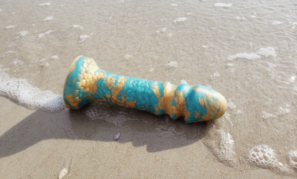 A large dildo laying on the shell-strewn sands of Daytona Beach. Water has glided across the sand to frame the dildo.