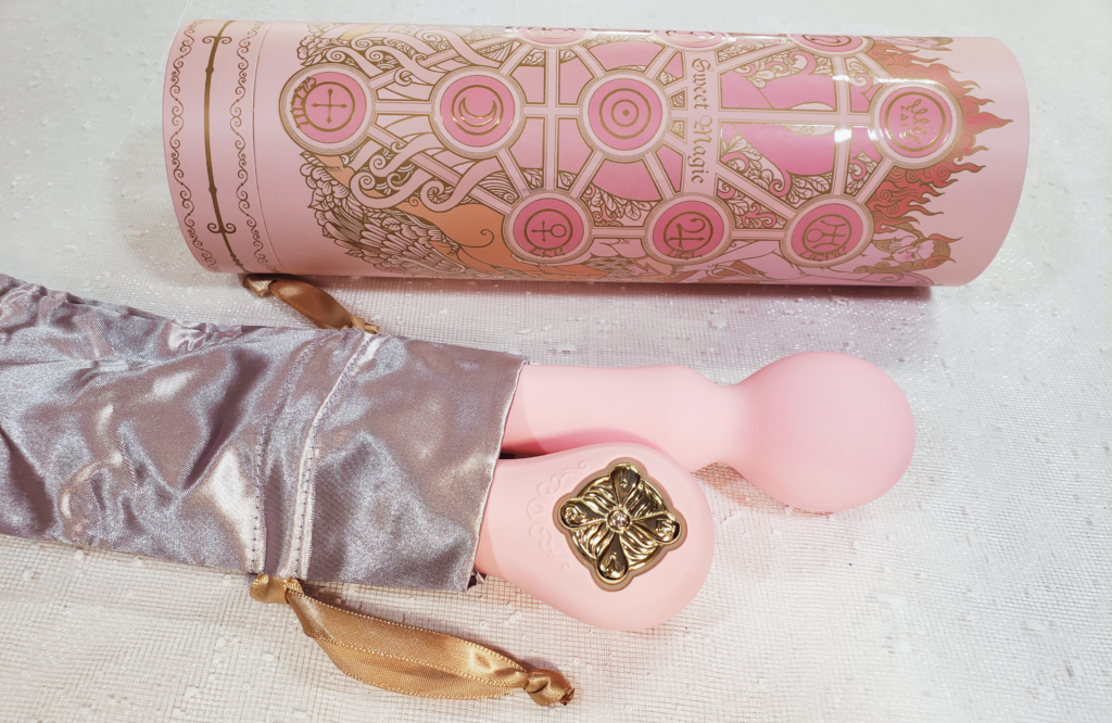 A tube-shaped box featuring a witchy, symbol-heavy design. Beside it, two pink sex toys are partially tucked inside a silver fabric bang with gold pullstrings.