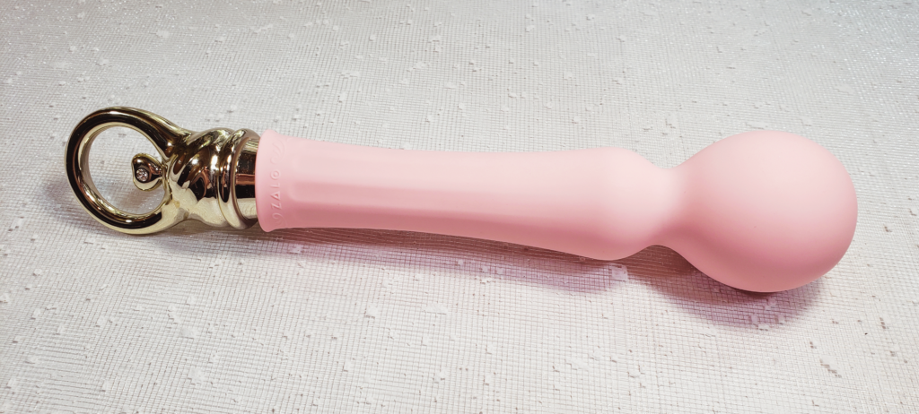  A pale pink vibrator shaped like a magic wand with a large orb at the end. Its base is a gold plastic loop, with a heart-shaped lever in the middle. It's laying on a white background flecked with snowflake-like texturing.