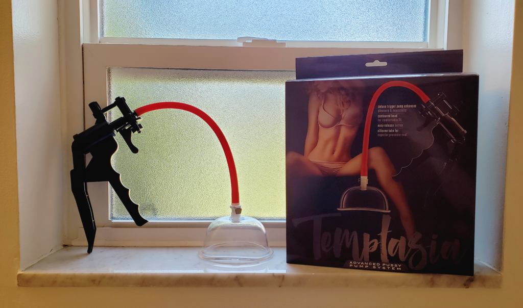 The Temptasia Pussy Pump propped up on a windowsill alongside its box. The window glass is textured for privacy. The pump consists of a large, black plastic handle and a translucent cup attached to a thin red tube. The box features a woman in her undies posed with legs spread.