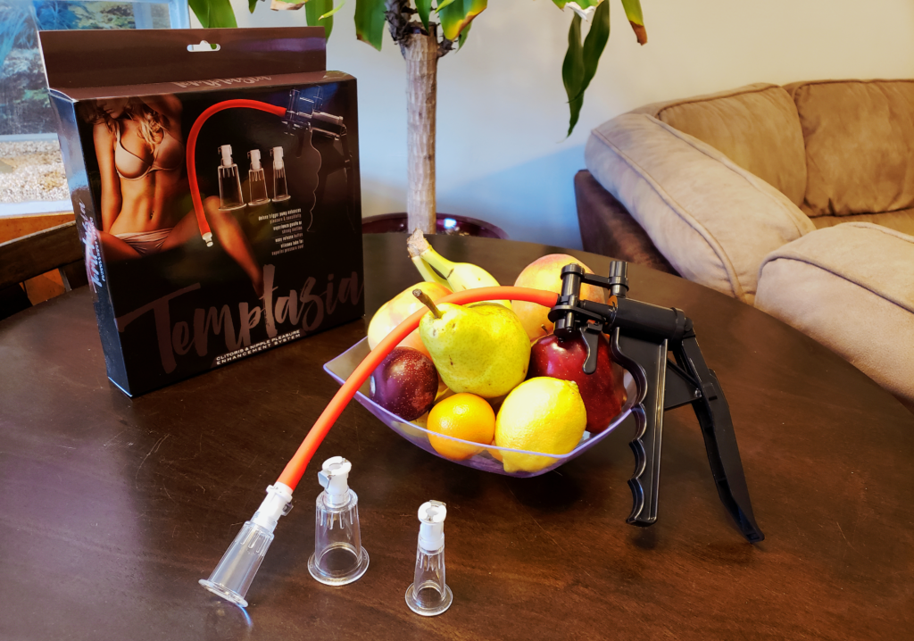 A clear bowl of fruit posed on a table. There is a plastic manual pump leaning against the bowl, and its packaging standing in the background. The packaging says Temptasia.