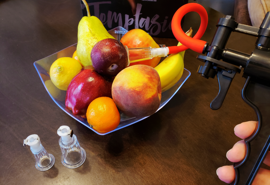 A white person's hand holding the handle of the pump, which is suctioned to a plum in the fruit bowl.