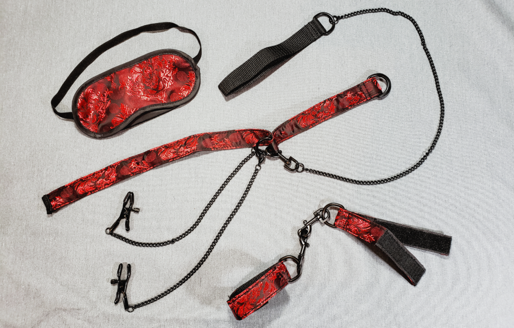 Bondage gear laying on a gray fabric background. There's a fabric blindfold, collar, and pair of cuffs. There is also a chain leash and clamps on chains.