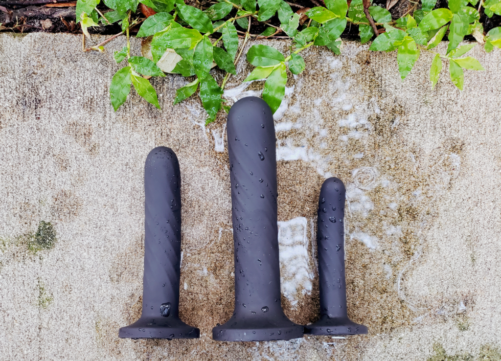 Three short, slim dildos in a charcoal black. They're laying in a reflective puddle beside dew-freckled foliage