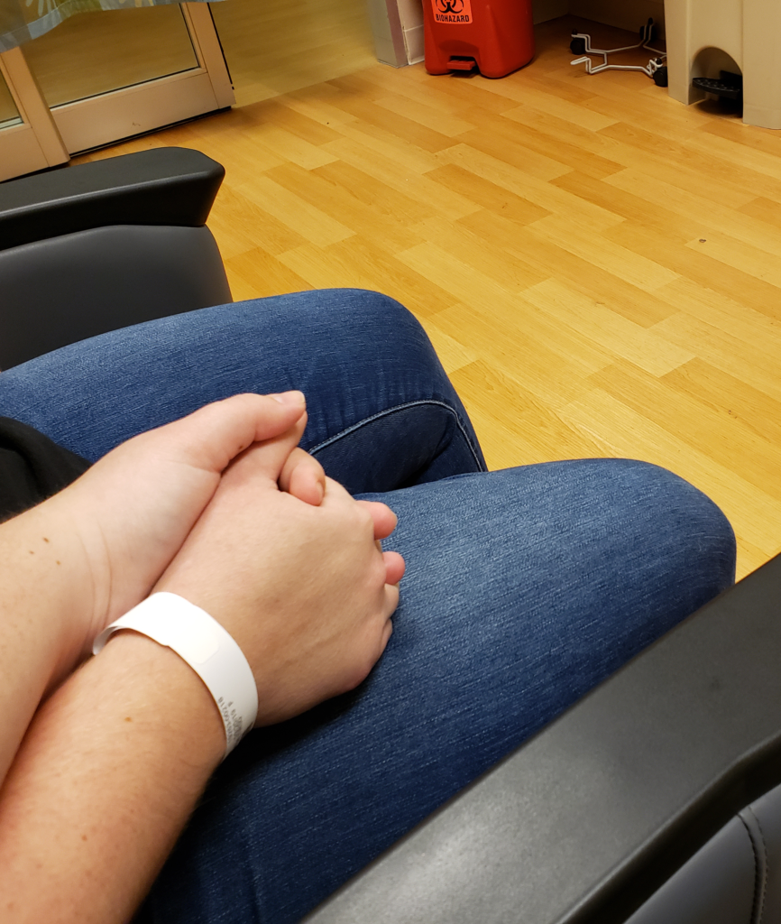 Two white nonbinary people's hands clutched on one person's lap. The edge of a hospital room door and a hazardous waste bin are visible in the corner.