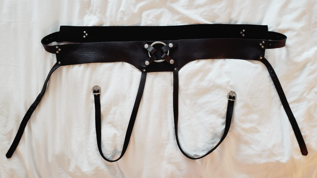 A black leather harness laying on rumpled white sheets.