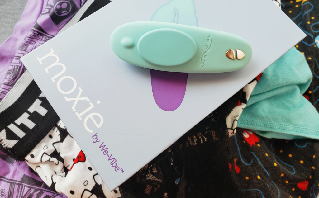 A small box sitting atop a pile of underwear in various styles. On the box - which says moxie - is a small, pastel blue-green vibrator with a magnetic attachment.