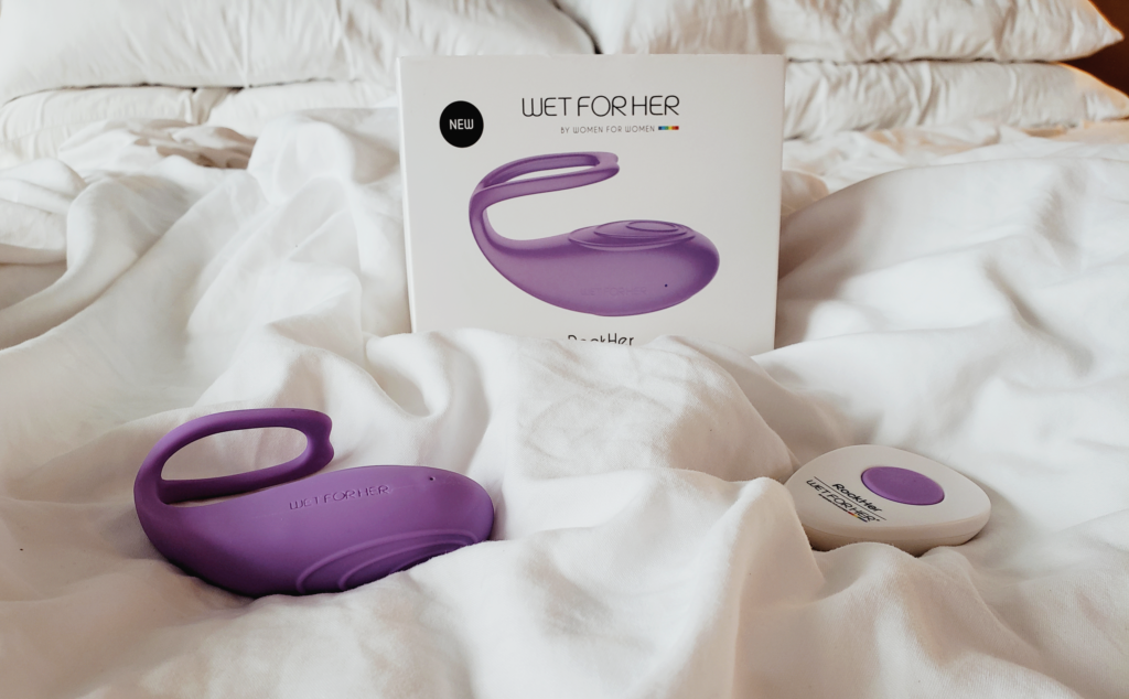 A sex toy box laying on a bed with white sheets. The box says WET FOR HER and features a curved toy that's almost whale shaped: a bulbous bottom and a tail-like smaller arm. The toy and its remote are laying on the sheets.