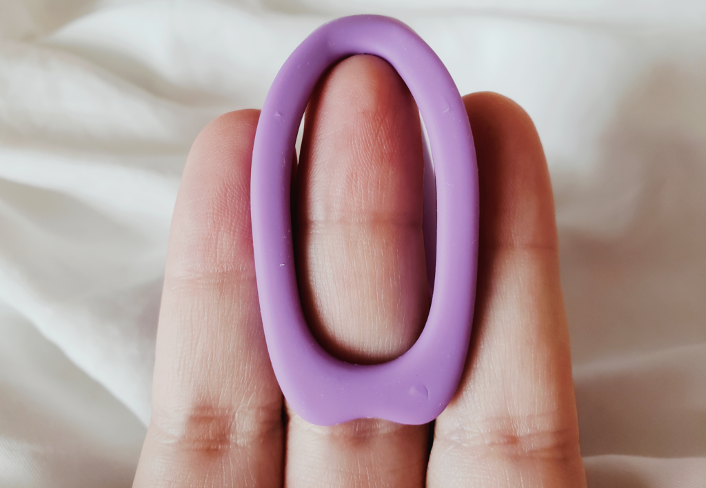 A white nonbinary person's fingers held behind the RockHer's smaller arm to demonstrate size. Their fingers are relatively large. There are some noticeable imperfections in the toy's silicone finish.