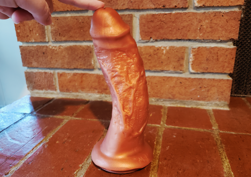 A bronze dildo sitting on a brick fireplace. A person's finger is posed on the toy's head to hold it up. Its shaft is detailed with unique dappling and divots.