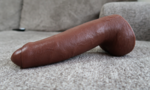 A realistic dildo in a dark skin tone, with foreskin and balls, laying on a gray couch.