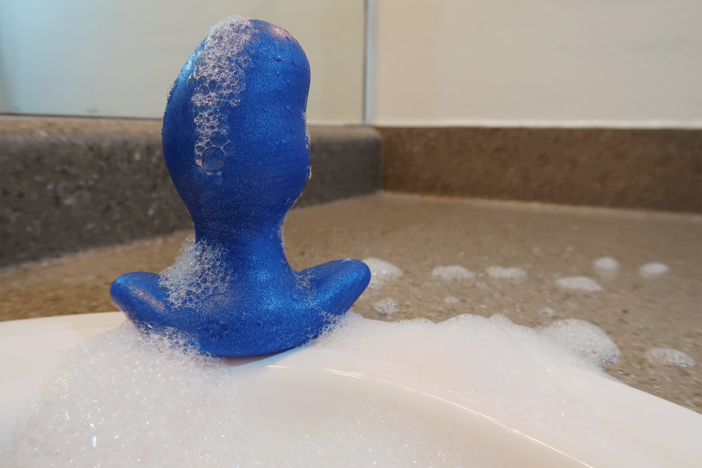 A picture of a large blue butt plug sitting on the edge of a sink, covered in suds. The plug is shaped like a kidney bean.