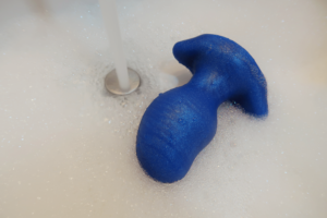 A blue butt plug laying in a sink with water spraying down beside it.