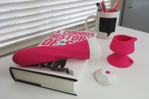 A fat pink vibrator laying on a copy of A Curious History of Sex. There's a mug with pink pens in it beside the vibe's detatched suction cup and remote.