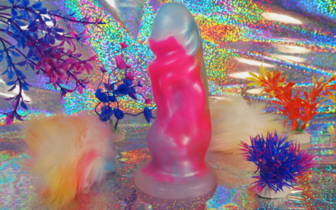 A thick, short dildo posed against a glittering background. It's surrounded by neon colored fake plants and yellow puff balls.