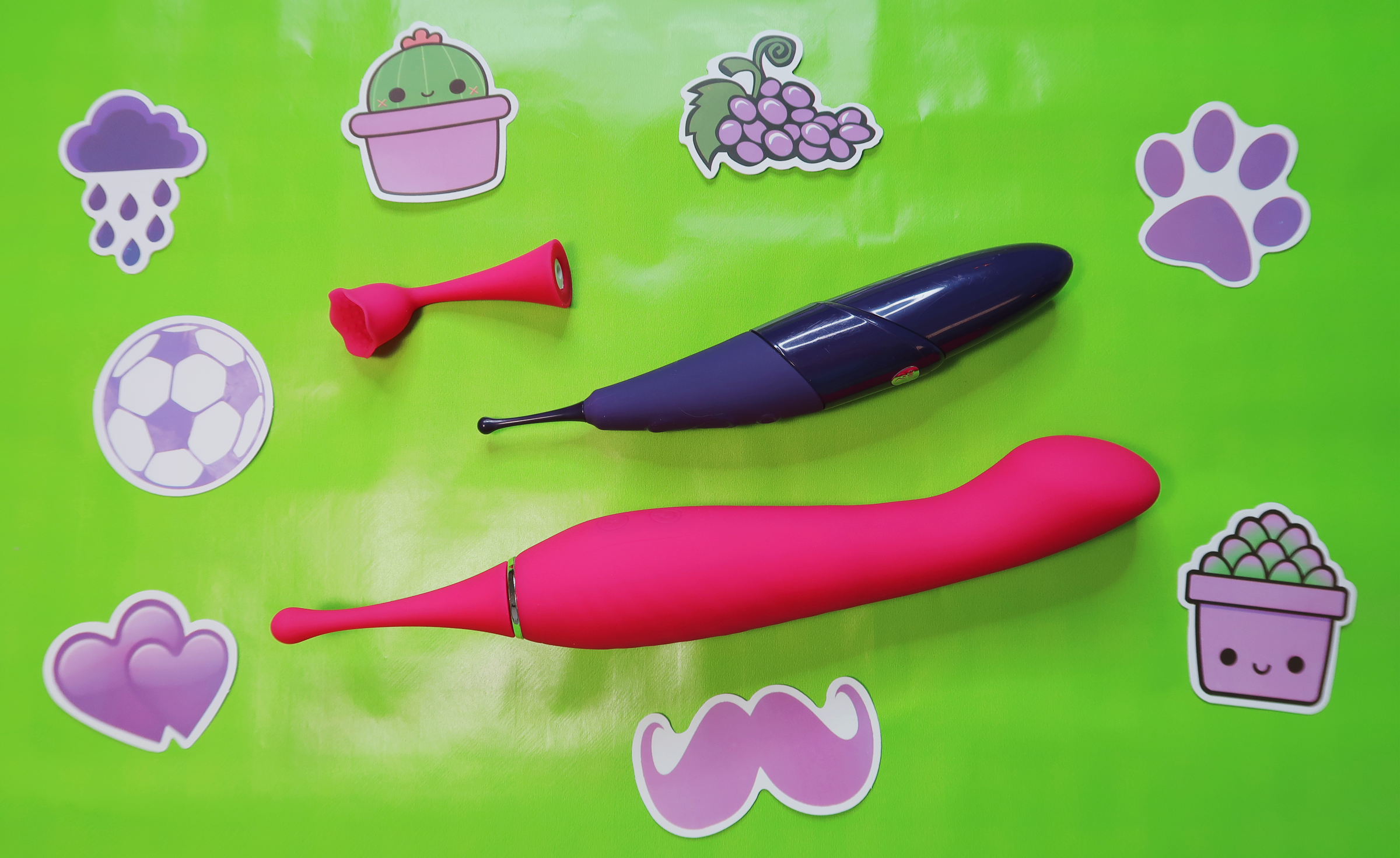 A Zumio laying on a green backdrop beside a larger, similarly pinpoint pink vibrator.