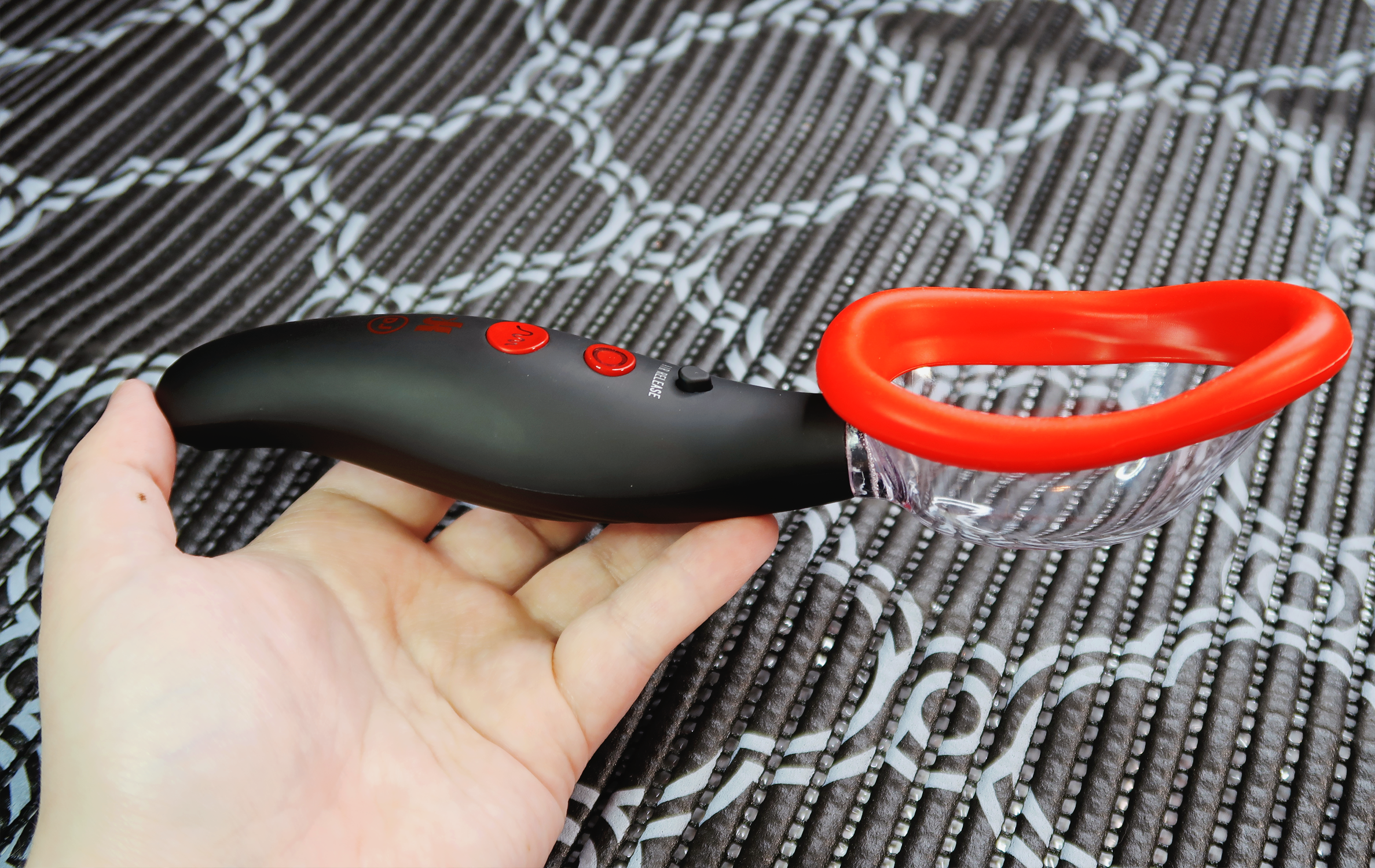 A white person's hand holding a spoon-shaped pussy pump. The pump is black with red buttons and trim.