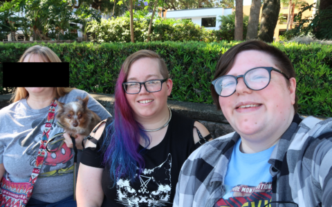 3 people sitting in a beautiful park. One person's face is censored out. The other two are fat, white, nonbinary folks in alternative clothes.