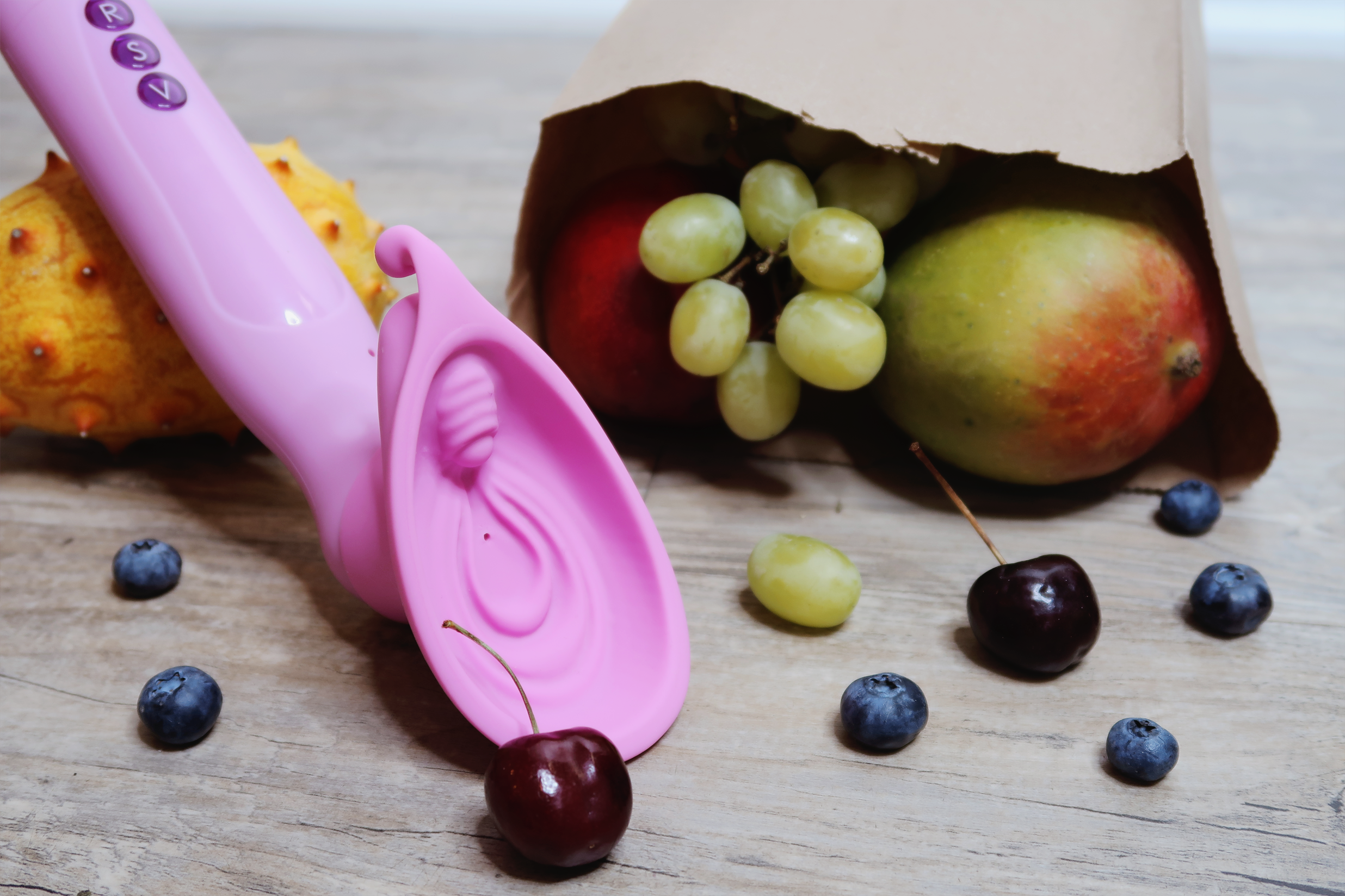 A light purple vibrator laying next to an open paper bag that has fruit spilling out. The vibrator has a vulva-shaped cup at the end that serves as a pump.