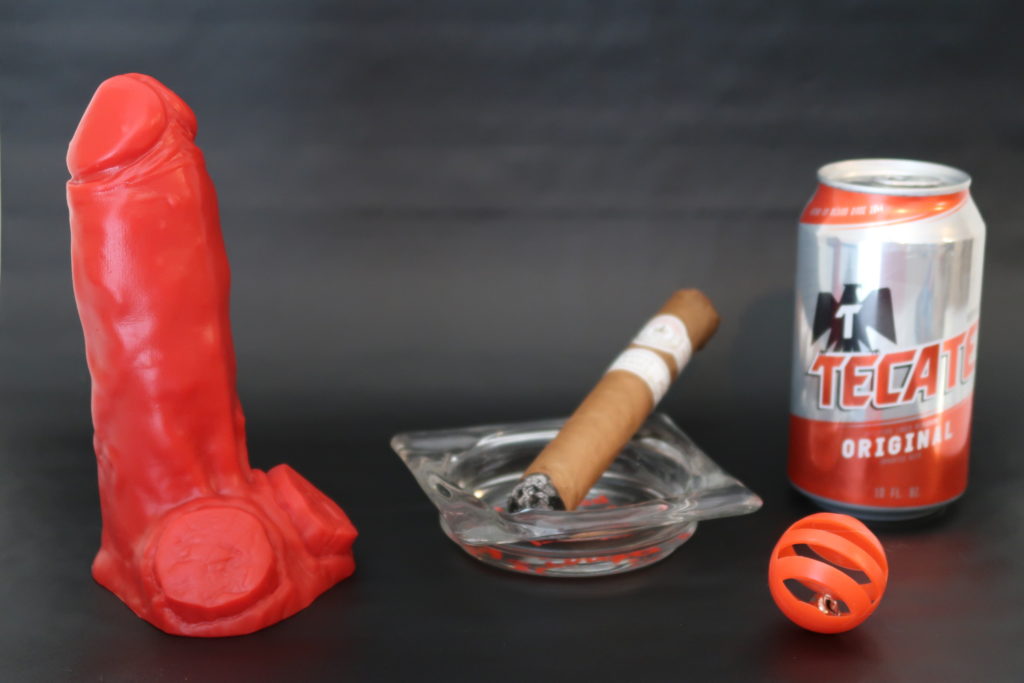 A thick red dildo with sawed-off horns for balls posed beside a cigar in an ashtray and a can of beer.