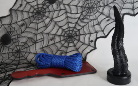 A lightly twisted black tongue dildo modeled after Venom's long tongue. It's posed in front of a spiderweb pattern, with a blue rope and red paddle laying beside it.
