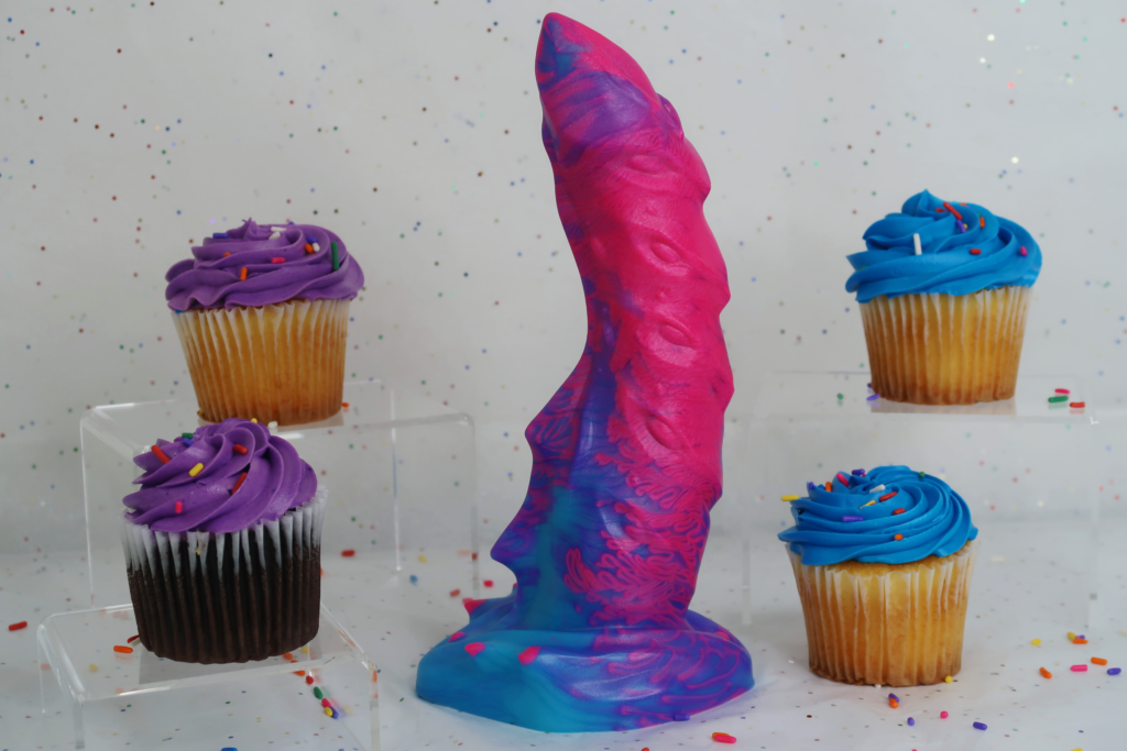 A neon-colored dragon themed dildo surrounded by cupcakes.