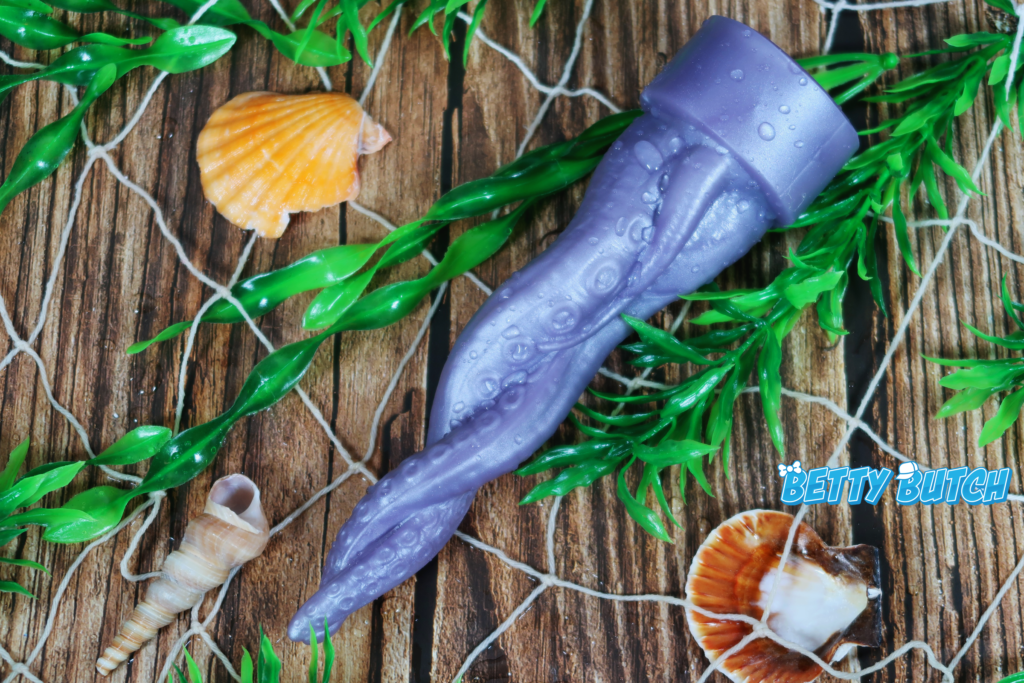 A light purple tentacle dildo laying on a fake wood background, surrounded by a damp net and plastic kelp.