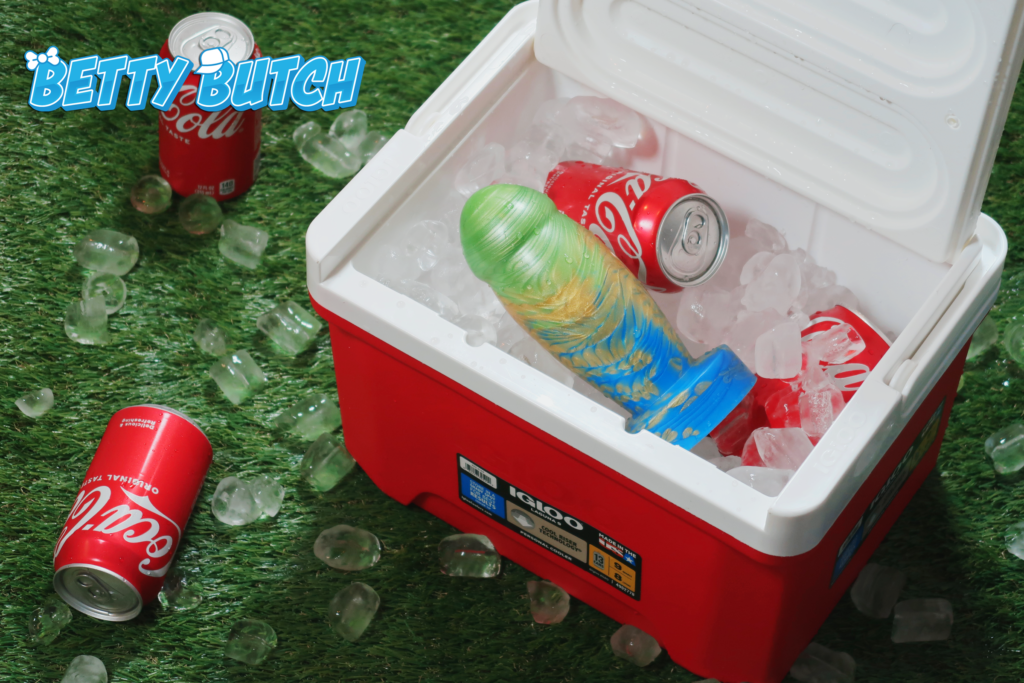 The Uberrime Amplus sitting in an open ice cooler alongside several cans of coke. There's chunks of ice scattered across the fake grass beneath.