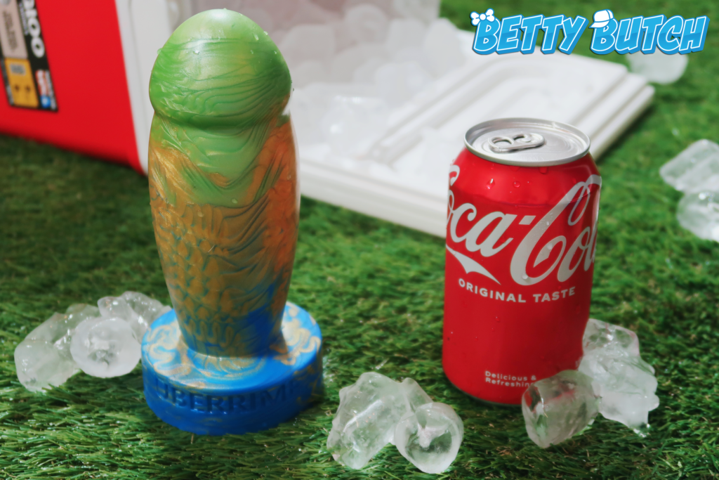 The Uberrime Amplus Abstract Dildo posed on fake grass with an ice cooler tipped over in the background. It's standing next to a can of coke for scale.