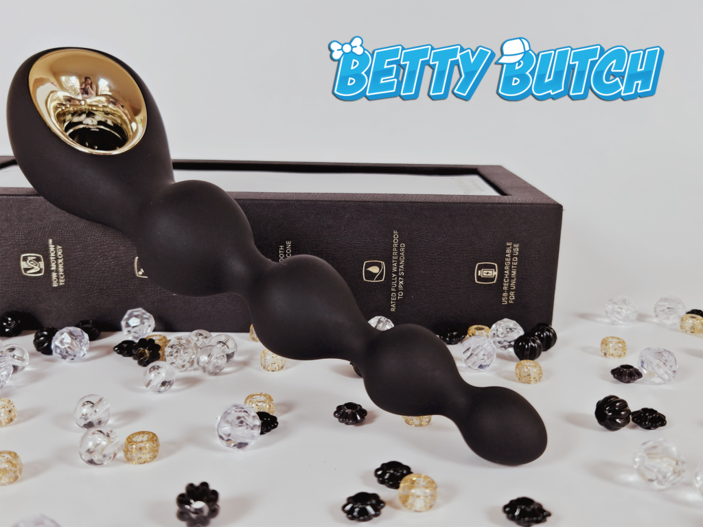 The Lelo Soraya Vibrating Anal Beads leaned up against their sleek, minimalist black packaging. There are beads scattered around the toy. 